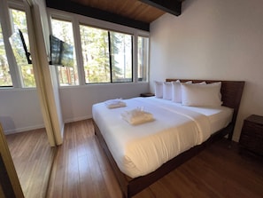 A bedroom with a wooden floor, a bed with white linens, and a window with a view of trees.