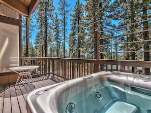 A hot tub on a wooden deck with a table and chairs, surrounded by tall pine trees.