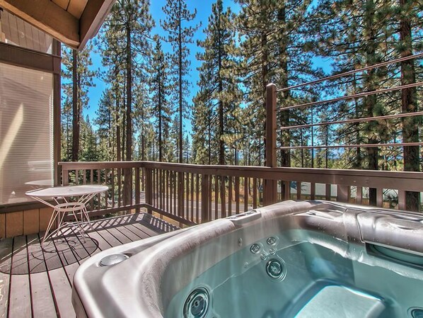 A hot tub on a wooden deck with a table and chairs, surrounded by tall pine trees.