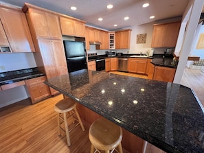 A modern kitchen with wooden cabinets, a black granite countertop, stainless steel appliances, and a bar stool.