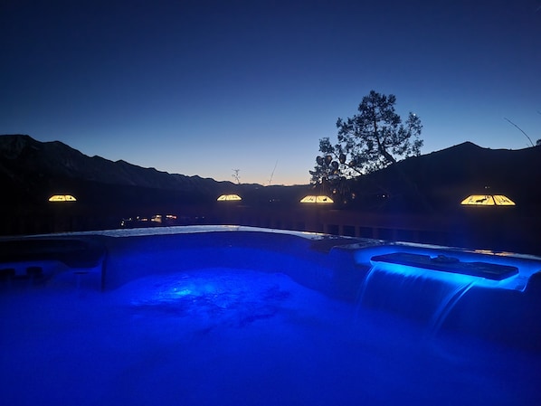 A hot tub area with a waterfall feature, surrounded by mountains and illuminated by warm lights.