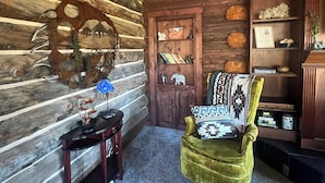Ein Blockhaus-Interieur mit Holzbalkendecke, rustikaler Wand, einem Holzstuhl, einem kleinen Tisch und einem Bücherregal mit verschiedenen Gegenständen.