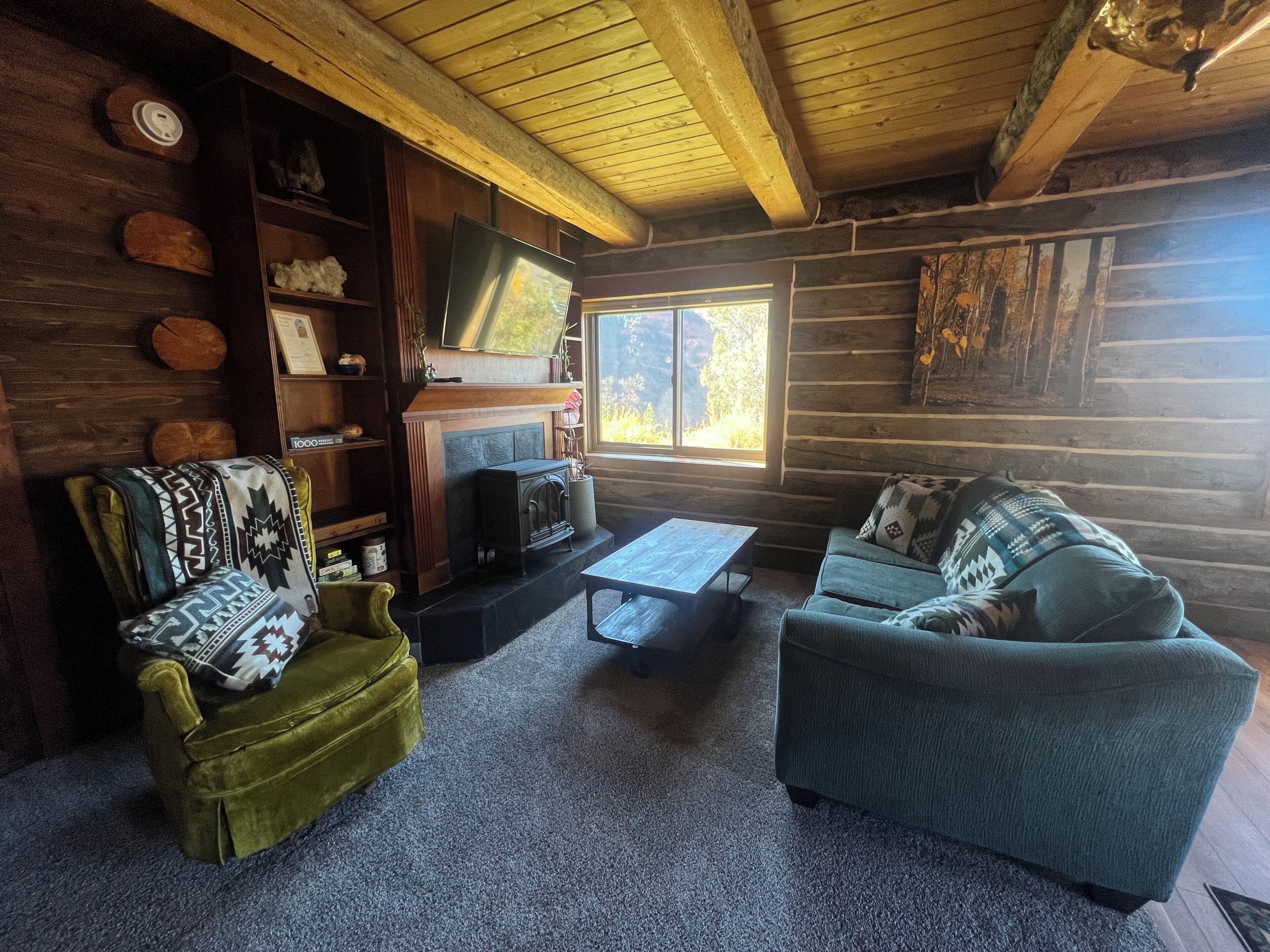 L’intérieur d’une cabane en rondins, avec une cheminée, un téléviseur, un fauteuil vert, un canapé bleu et une grande fenêtre.