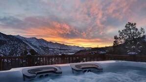 Ein Whirlpool mit Holzterrasse, umgeben von schneebedeckten Bergen und einem Sonnenuntergangshimmel.