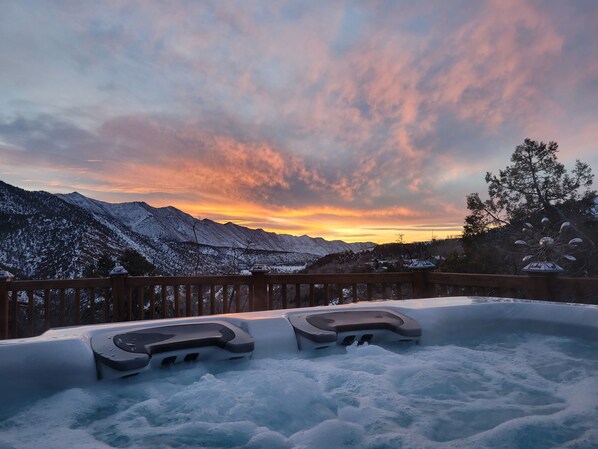 A hot tub with a wooden deck, surrounded by snow-covered mountains and a sunset sky.