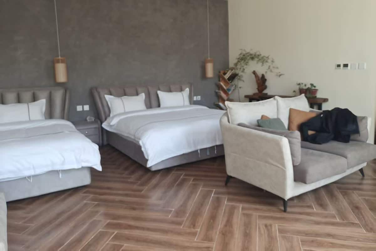 A modern hotel room with two beds, a sofa, and a wooden herringbone floor.