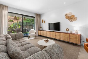Ein modernes Wohnzimmer mit einer großen Couch, einem Fernseher und einem Essbereich, der durch eine Glastür sichtbar ist.