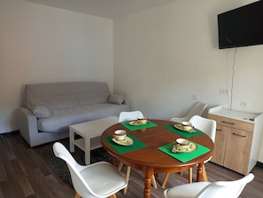 Ein Essbereich mit einem runden Holztisch, vier weißen Stühlen, einem grauen Sofa, einem weißen Schrank und einem an der Wand befestigten Flachbildfernseher.