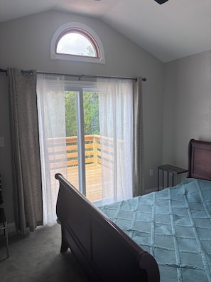 A bedroom with a bed, a window with curtains, a small table, and a view of a wooden deck.