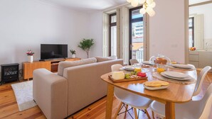 Un salon moderne avec un canapé, un téléviseur, une table à manger dressée pour le petit-déjeuner et une grande fenêtre.