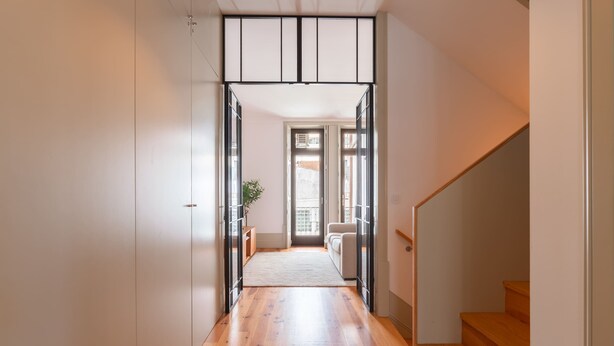 Un couloir au plancher en bois, un escalier sur la droite et une porte donnant sur une pièce avec une fenêtre.