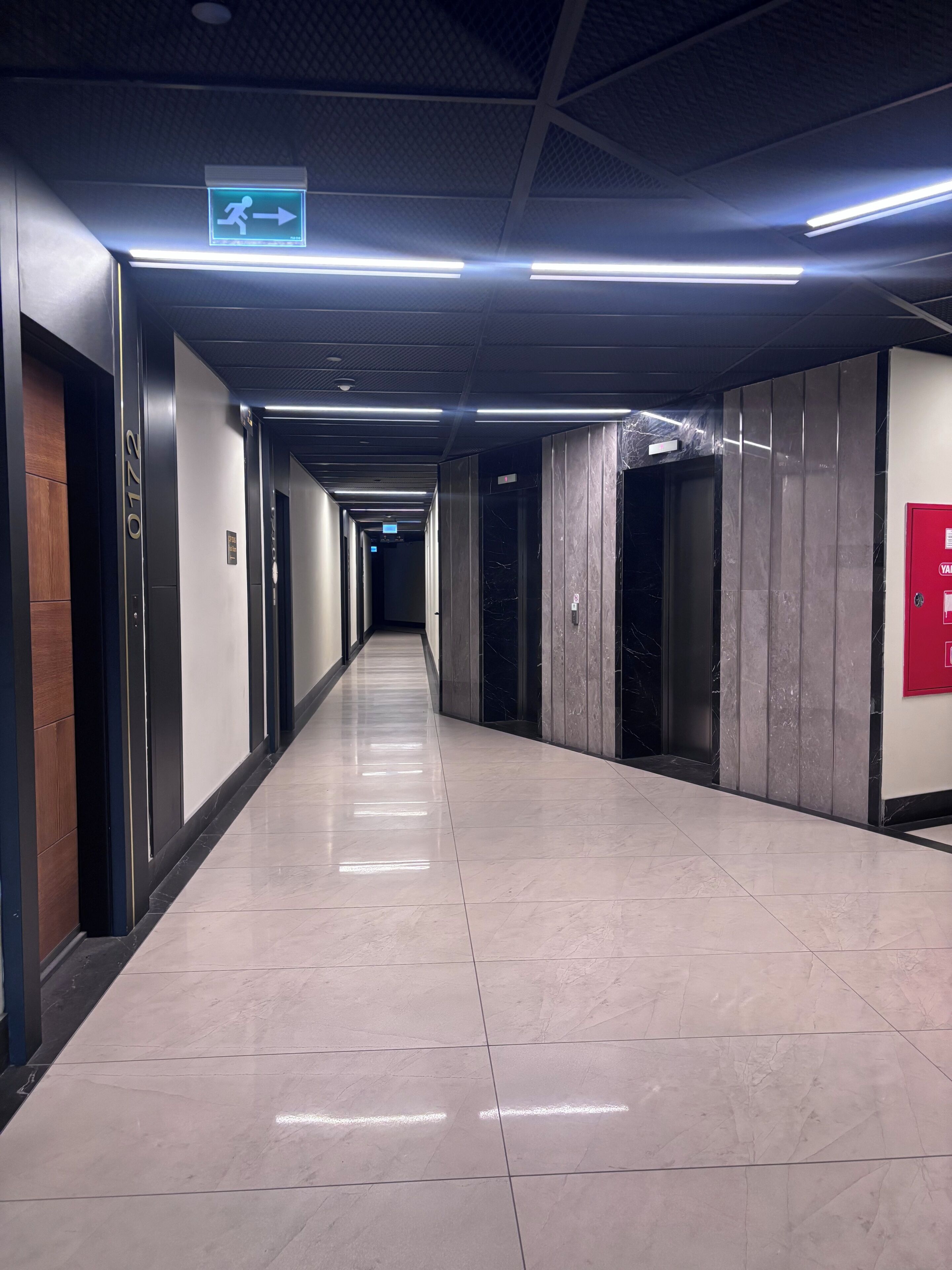 A long, well-lit corridor with a directional sign, elevators, and a red emergency exit sign.