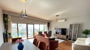 A modern living room with a dining area, a sofa set, a TV, and a large window offering a city view. - Benesta Centro by GOLDFINCH (Istanbul)