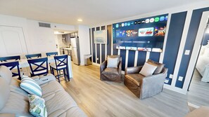A modern living room with a sofa, chairs, a TV, and a kitchen in the background. - New Luxurious 1 BR/2 Ba with 2 Kings and a Queen w/ Free Beach Service for 4! (Fort Walton Beach)