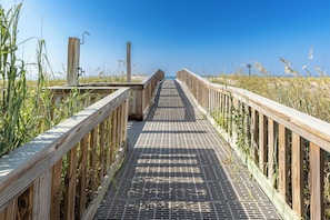 Une promenade en bois menant à une plage, entourée de hautes herbes et bordée d’un ciel bleu clair.