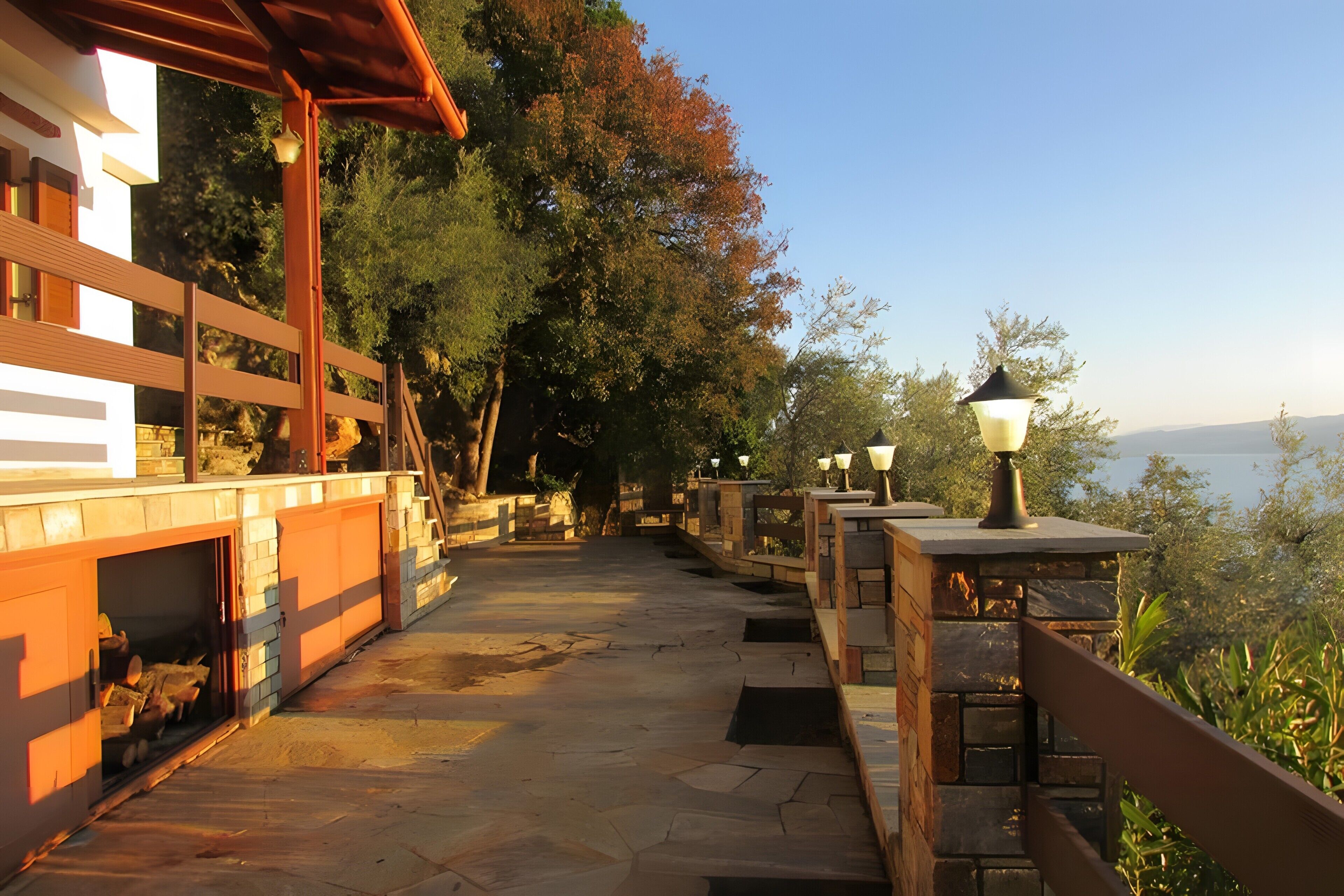 A stone pathway leading to a wooden deck with a railing, a stone wall with a lantern, and a view of the water.