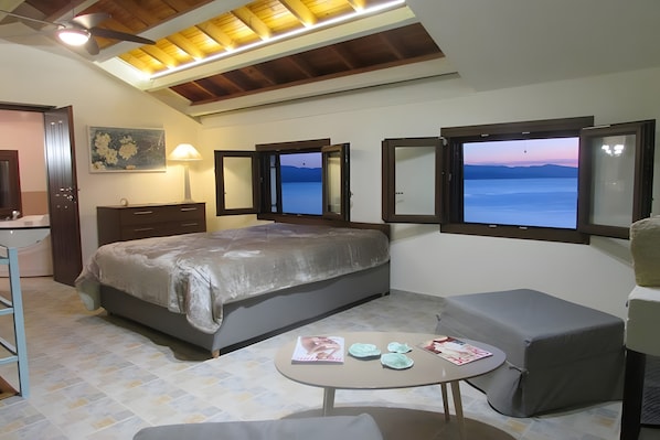 Un dormitorio con cama, escritorio, silla, mesa con revistas y vistas al mar a través de una ventana.