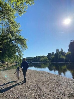 Ein Flussufer mit einer Brücke im Hintergrund, ein klarer Himmel und eine Person, die mit einem Kind spazieren geht.