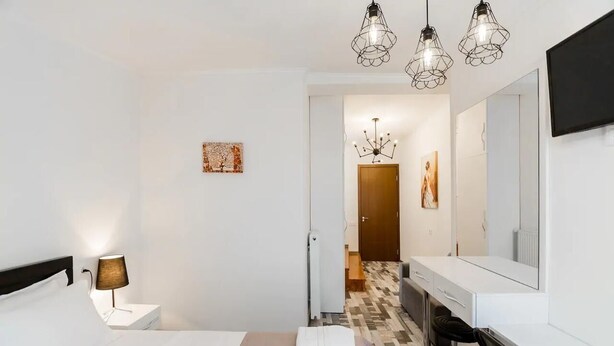 Hotellihuoneessa on sänky, yöpöytä, lamppu ja suuri peili. Huoneesta on ovi, joka johtaa käytävään, jossa on tuoli ja taulu.