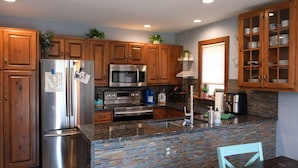 A modern kitchen with wooden cabinets, stainless steel appliances, a granite countertop, and a dining area with a chair.