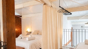 A bedroom with a wooden floor, a bed with white bedding, a bedside table, a hanging light, and a window with curtains.