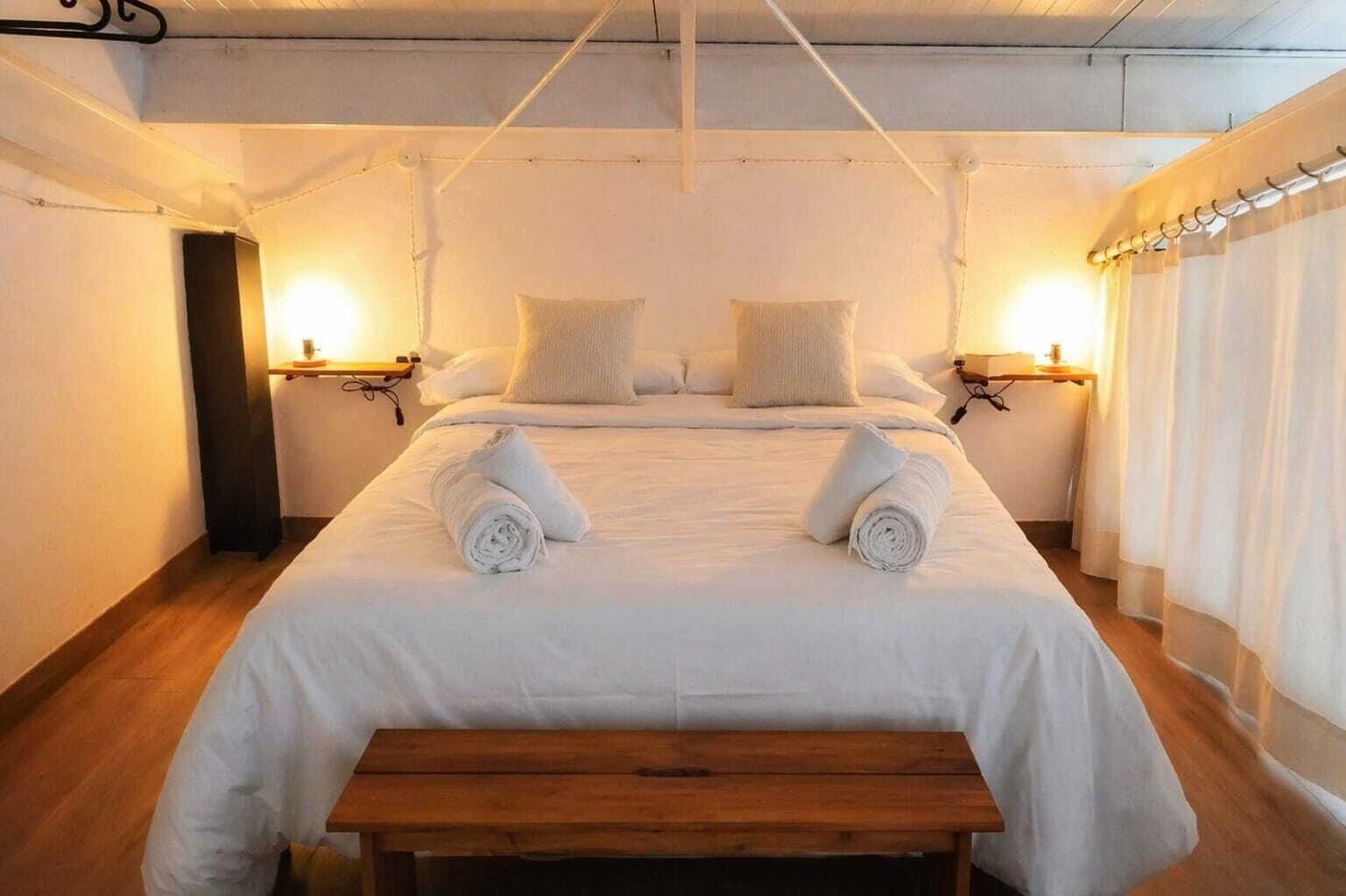 Habitación de hotel con una cama grande, dos mesitas de noche con lámparas, un banco de madera y una cortina en la ventana.