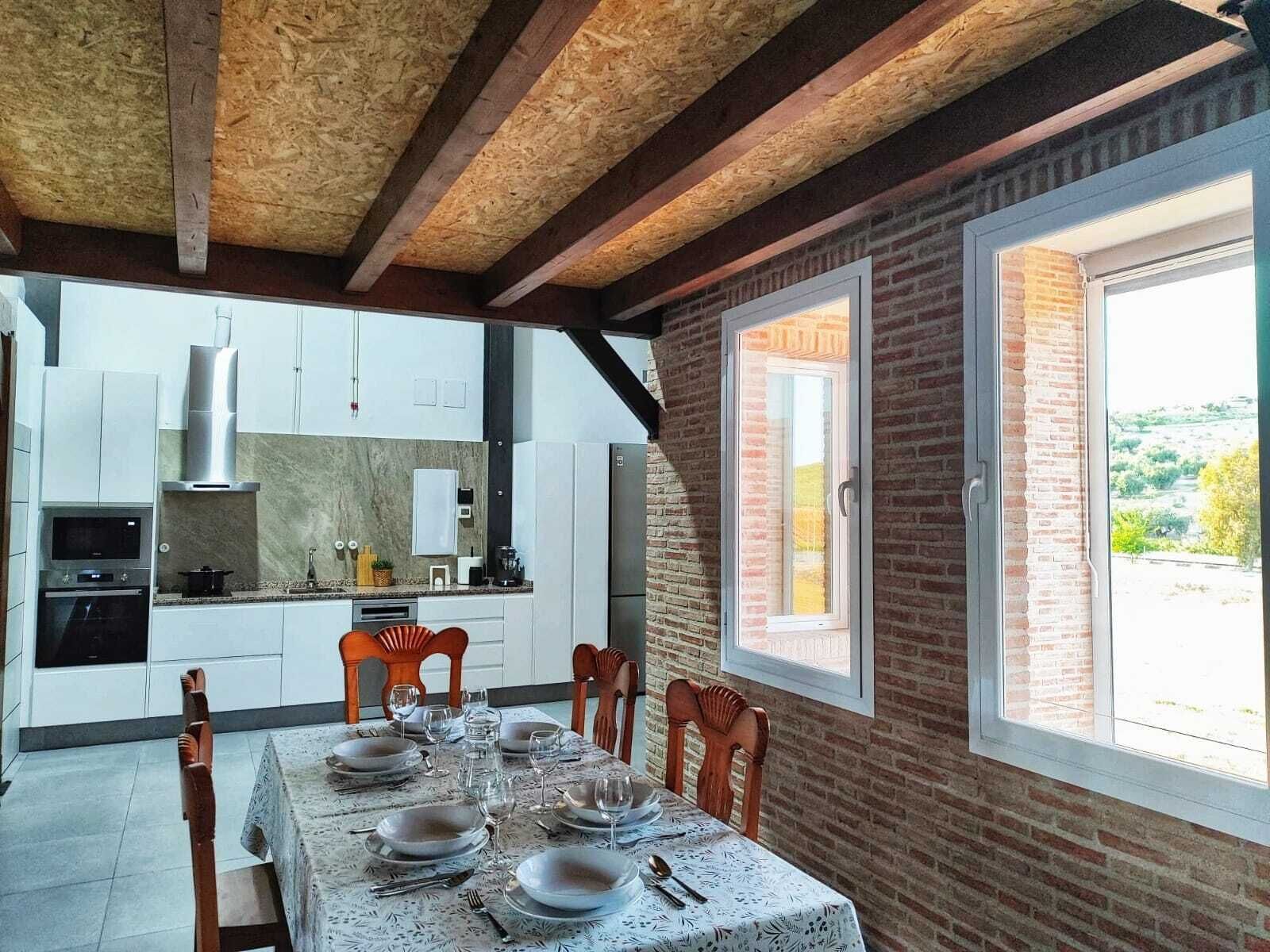 Uma área de refeições com mesa posta para quatro pessoas, uma cozinha com eletrodomésticos modernos e uma parede de tijolos com uma janela.