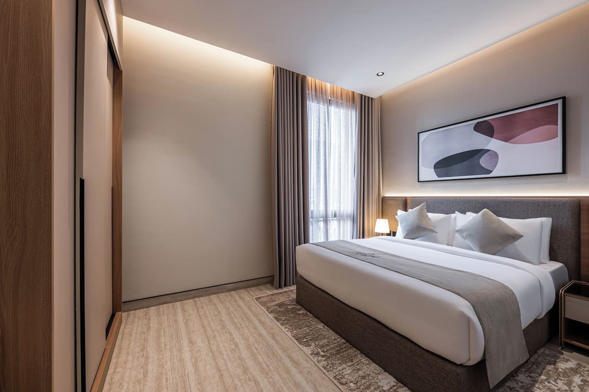 Habitació d'hotel moderna amb un llit gran, una tauleta de nit, un quadre abstracte penjat a la paret i una finestra gran amb cortines.