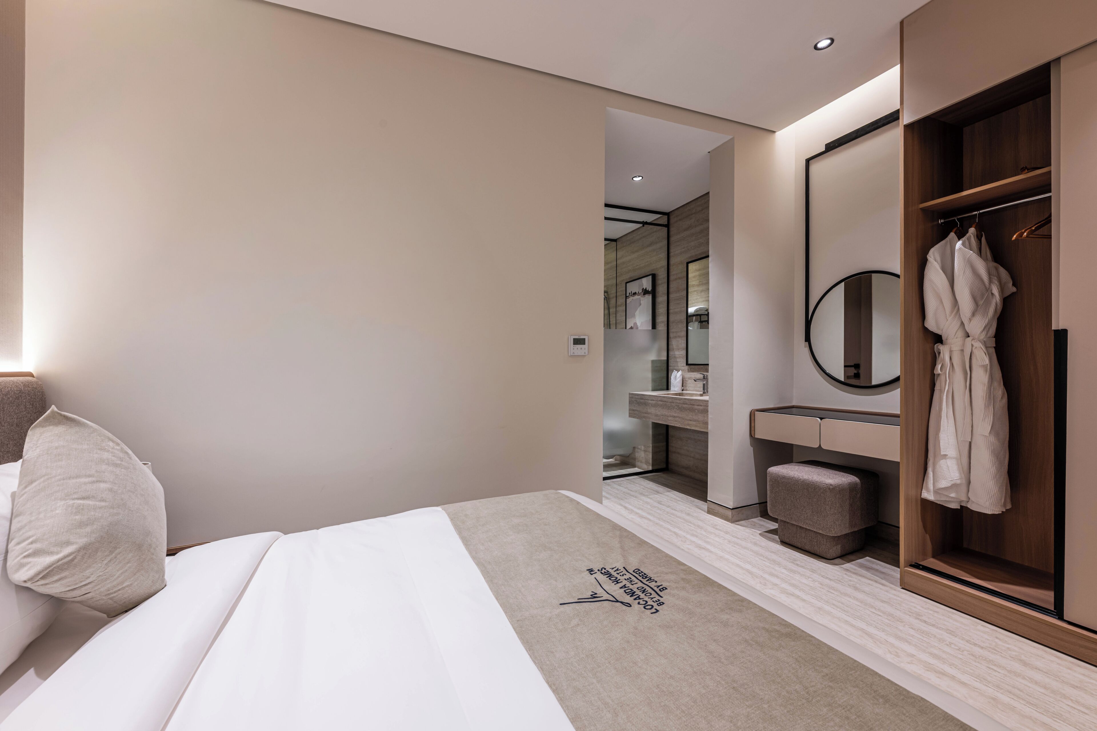 A modern hotel room with a bed, a robe hanging on a wooden closet, a round mirror, and a small stool.