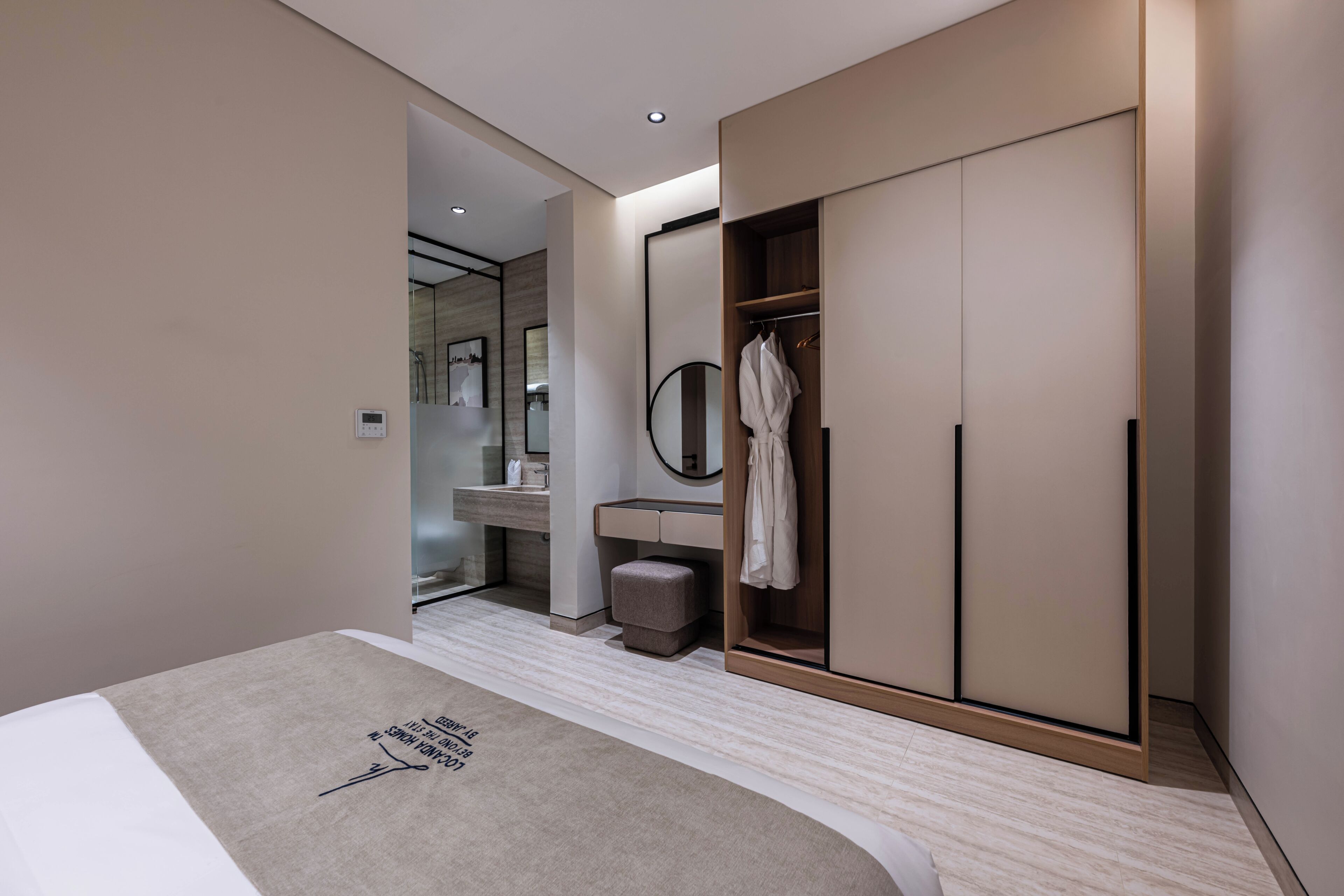 A modern hotel room with a bed, a walk-in closet, a bathroom with a mirror, and a small stool.