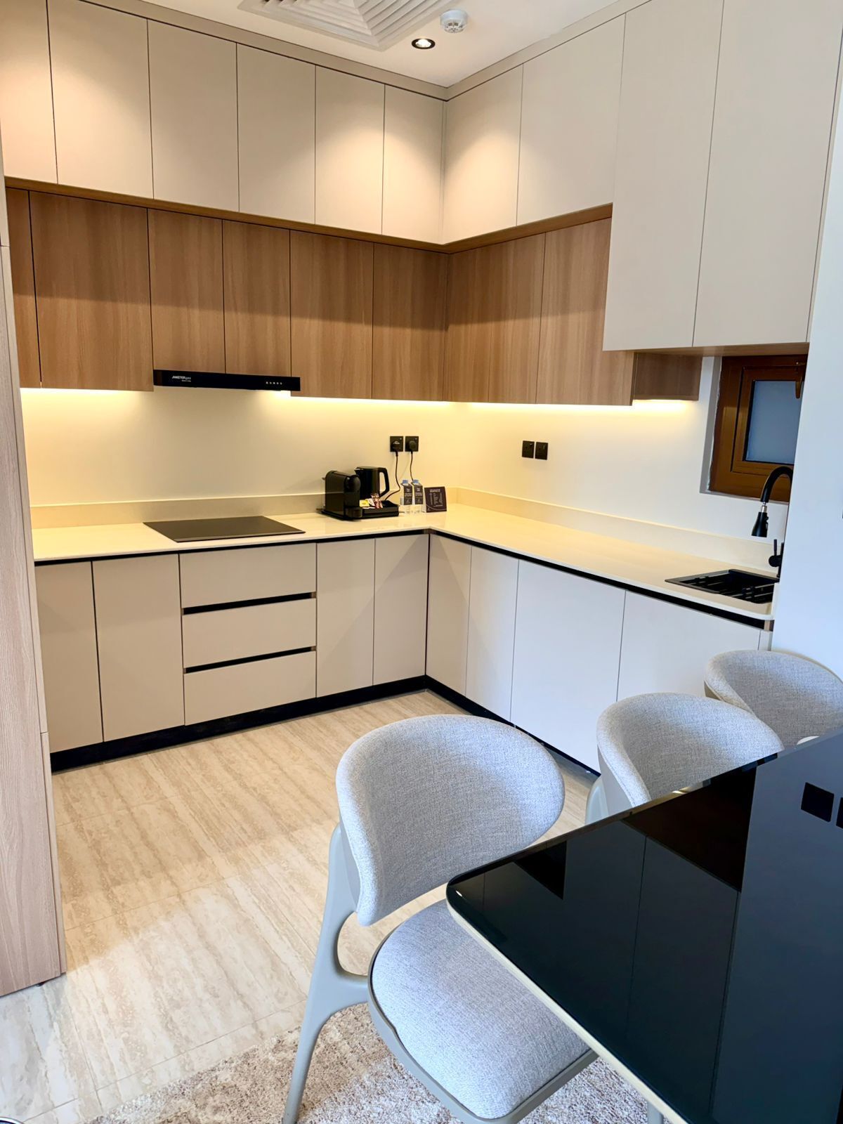 Una cocina moderna con estufa integrada, cafetera y un lavamanos pequeño. En primer plano, hay dos sillas grises y una mesa negra.