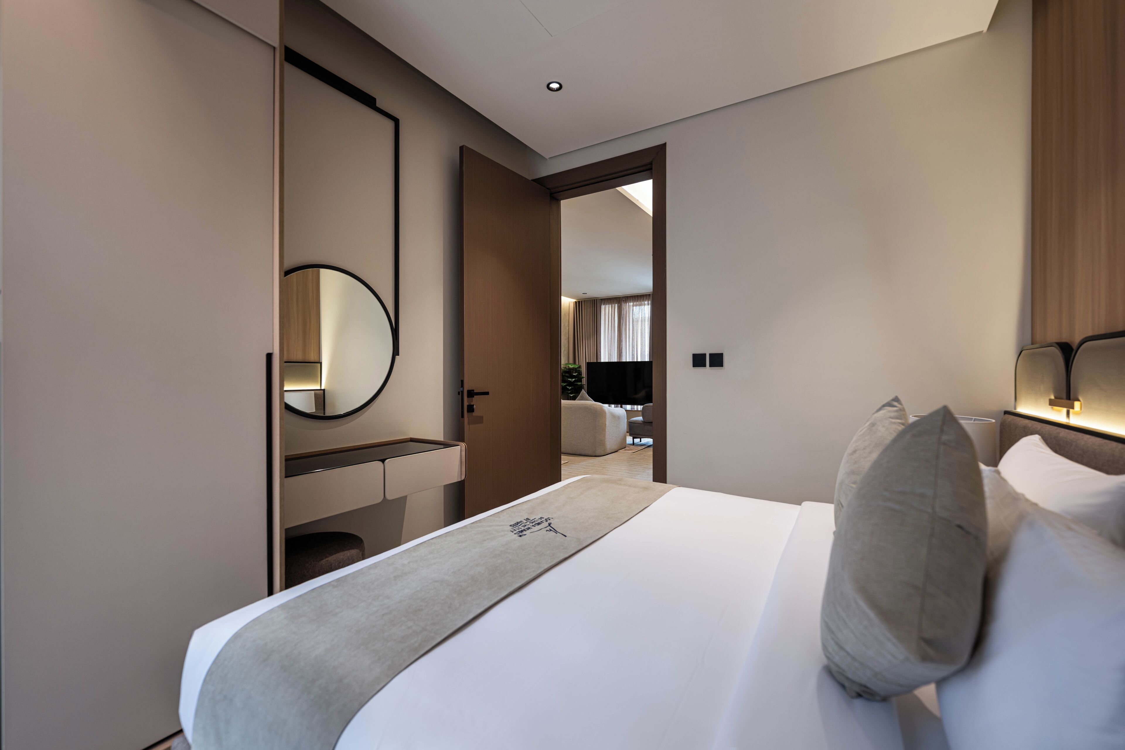 A hotel room with a bed, a bedside table, a mirror, a door leading to another room, and a wall-mounted light fixture.