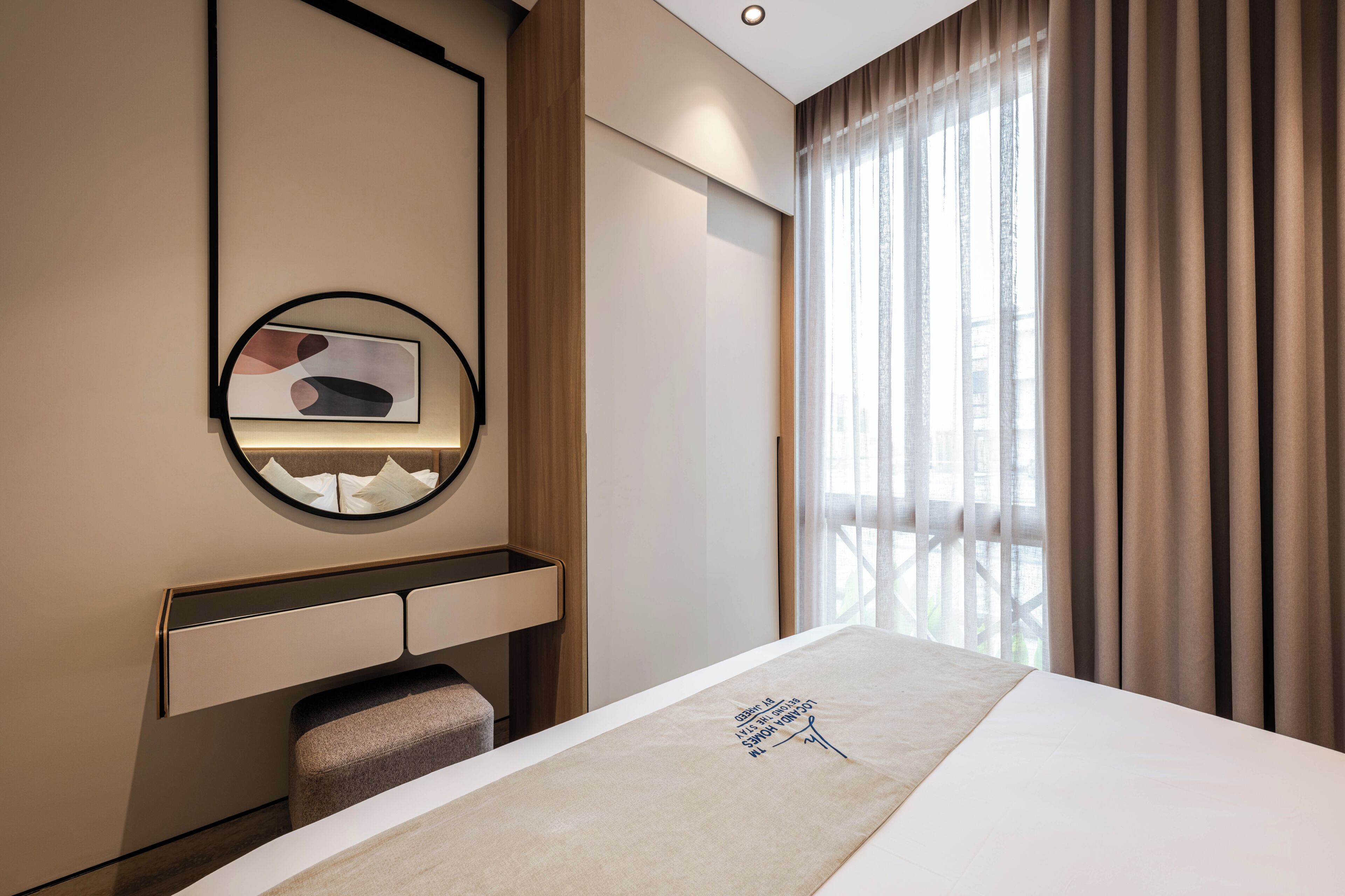 A hotel room with a bed, a bedside table, a mirror, a window with curtains, and a wall-mounted artwork.