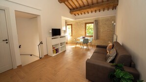 A living room with a brown sofa, a TV, a dining table with chairs, and a brick wall. - Beautiful renovated farmhouse in the relaxing Umbrian countryside (Sant'Angelo di Celle)
