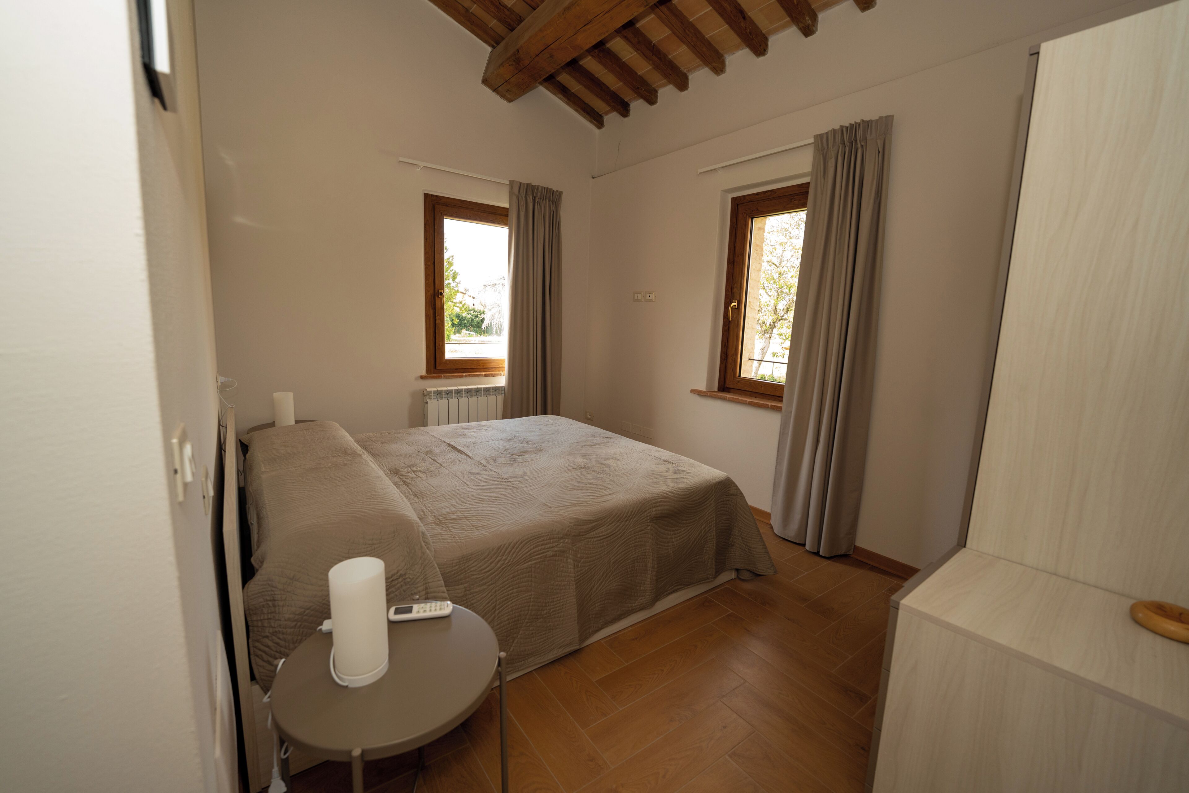 Une chambre avec un lit, une table ronde sur laquelle se trouve une télécommande, deux fenêtres avec des rideaux et un plafond en bois.