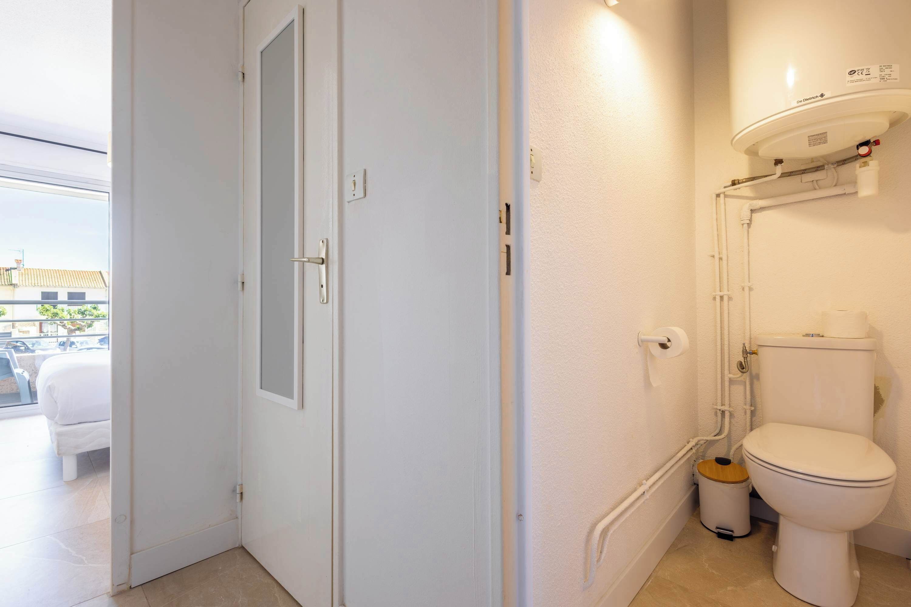 A bathroom with a toilet, a small trash bin, and a window leading to a balcony.