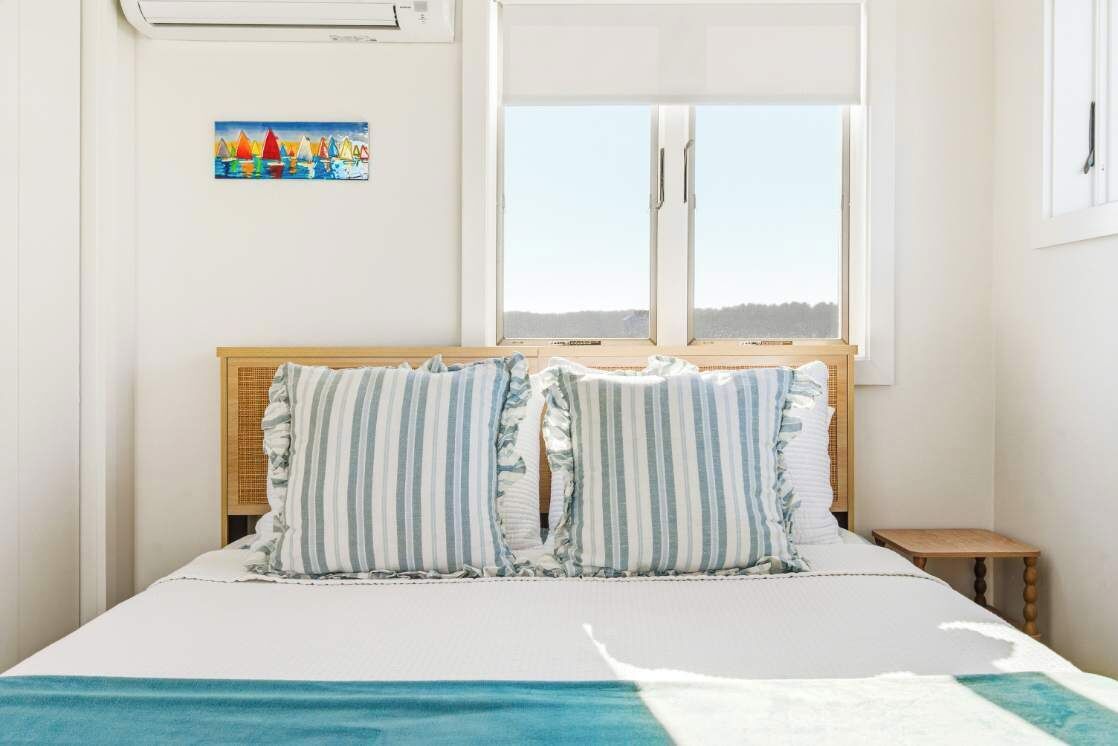 Un dormitorio con cama, ventana, una mesita y aire acondicionado instalado en la pared.