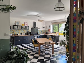Eine Küche mit Schachbrettboden, einem Esstisch mit Stühlen, einem Deckenventilator und einem Fenster mit Blick ins Grüne.