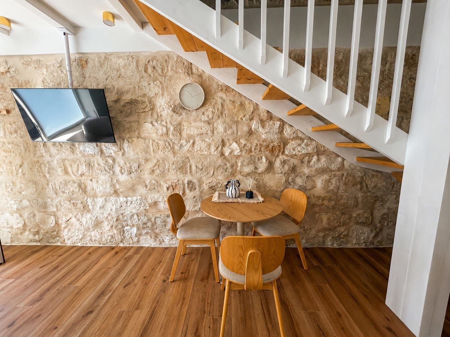 A room with a stone wall, a wooden floor, a round table with chairs, a flat-screen TV mounted on the wall, and a white staircase.