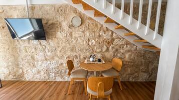 A room with a stone wall, a wooden floor, a round table with chairs, a flat-screen TV mounted on the wall, and a white staircase.