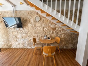 Ein Zimmer mit einer Steinwand, Holzboden, einem runden Tisch mit Stühlen, einem an der Wand befestigten Flachbildfernseher und einer weißen Treppe.
