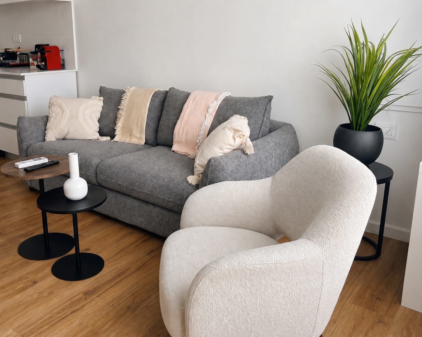 A modern living room with a grey sofa, a white armchair, a small round table with a white vase, and a potted plant.