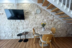 A room with a stone wall, a flat-screen TV mounted on it, a dining table with chairs, a small round table with a phone, and a staircase with wooden steps. - לופט אורבני ליד המושבה הגרמנת (Haifa)