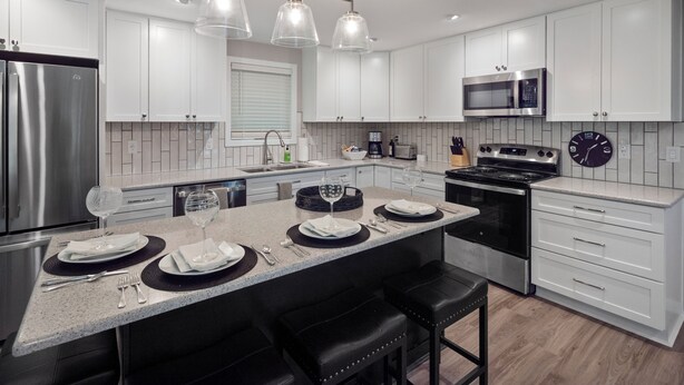 A modern kitchen with white cabinetry, stainless steel appliances, a central island with bar stools, pendant lighting, and a microwave above the oven.