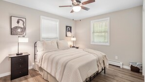 A bedroom with a bed, a ceiling fan, two bedside lamps, a nightstand, a dresser, and two windows with blinds.