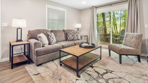 A living room with a sofa, coffee table, lamps, and a view of trees through a sliding door.