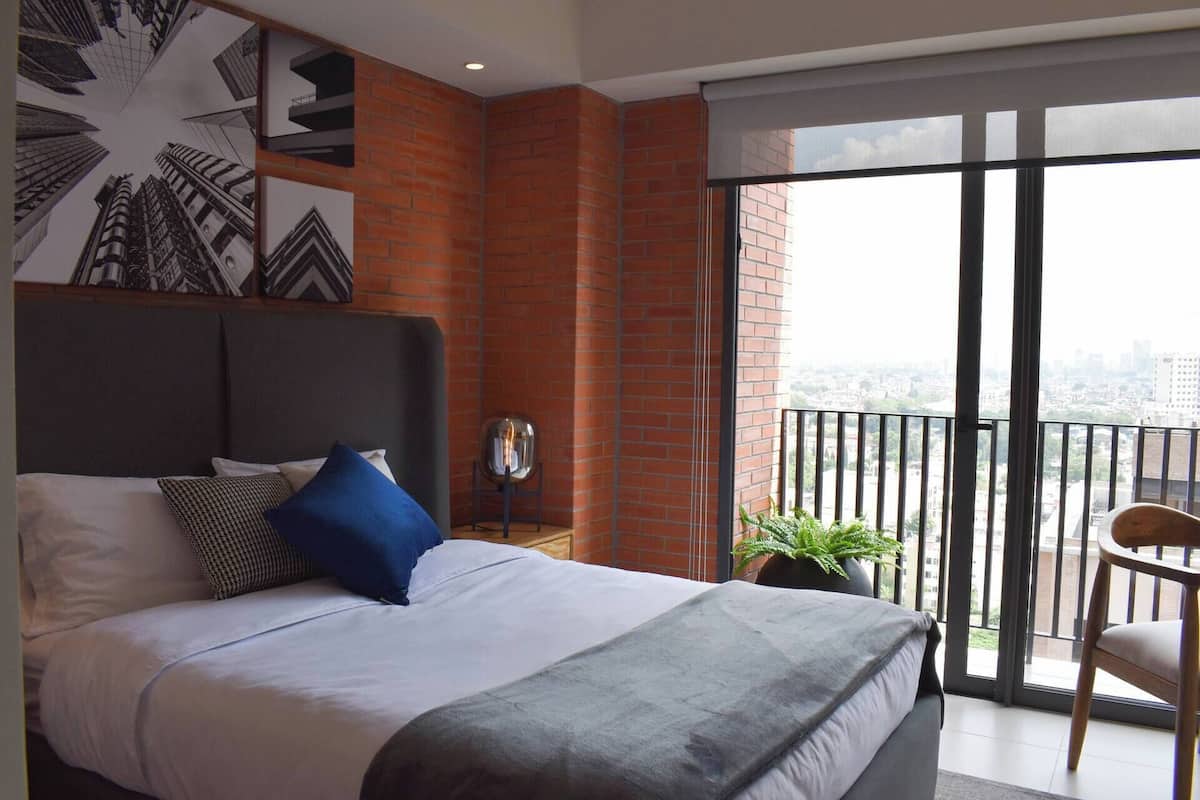 A bedroom with a large bed, a view of the city through a balcony, and a brick wall.