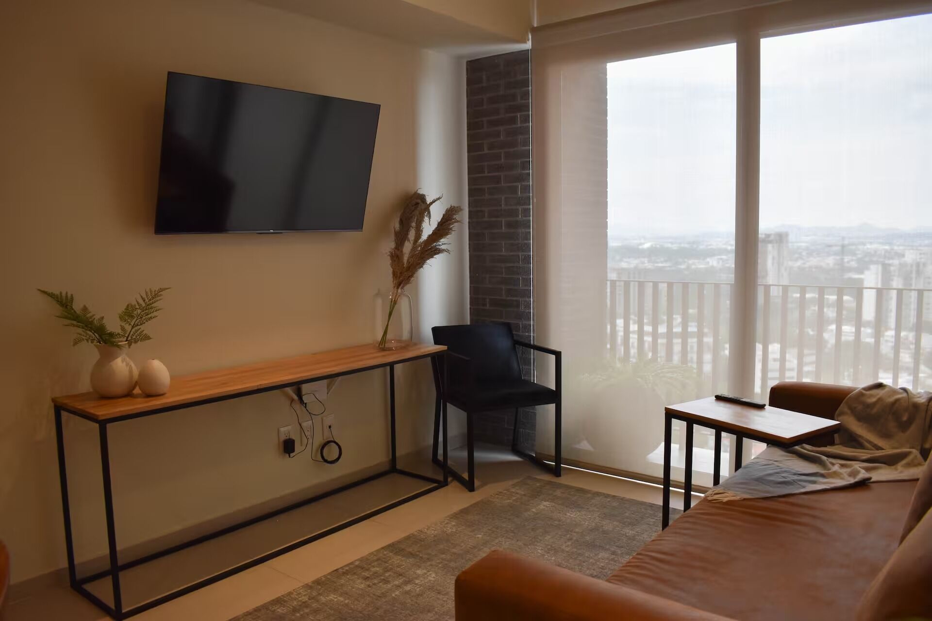 A modern living room with a TV, a black chair, a wooden console table, and a view of the city through large windows.