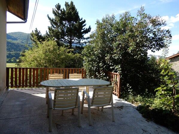 An outdoor patio with a round table and two chairs, surrounded by a wooden fence, overlooking a lush green landscape with trees and hills.