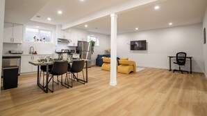 Una cocina moderna con armarios blancos, un comedor con mesa y sillas, y una sala de estar con sofá y televisor en la pared.